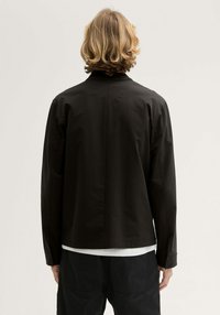 Czarna kurtka z długimi rękawami i prostym kołnierzykiem, charakteryzująca się prostym krojem pleców i lekkim materiałem. Nosi się ją na białej koszuli.