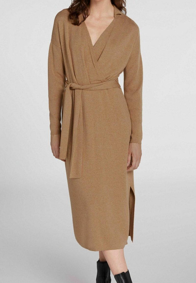 Femme portant une robe portefeuille camel à manches longues avec ceinture à la taille et fente latérale, associée à des bottines noires.