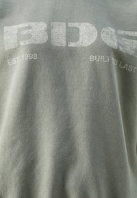 Melanžové šedé tričko vyrobené z bavlny, s poškozeným potiskem obsahující "BDG", "EST 1998" a "BUILT TO LAST" v bílém písmu.