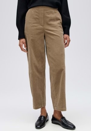 Persona che indossa pantaloni in velluto beige a vita alta e mocassini neri, in piedi con le mani rilassate lungo i fianchi davanti a uno sfondo semplice.