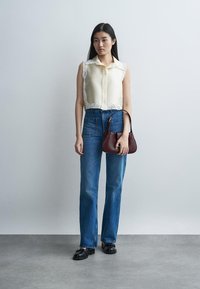 Jauna moteris, vilkinti smėlio spalvos be rankovių palaidinę su nėrinių apdaila, mėlynais aukštos liemens džinsais, juodais loaferiais, laikanti mažą rudą rankinę, stovinti patalpose.