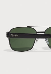 Ochelari de soare Ray-Ban cu o ramă metalică neagră, lentile verzi, formă rectangulară și un logo distinctiv pe lateral. Inclusiv suporturi pentru nas transparente.
