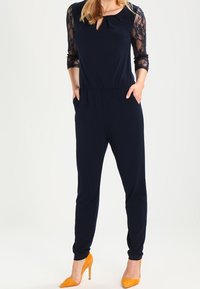 Kvinna bär en marinblå jumpsuit med spets ärmar i tre kvartar och fickor, ihop med klarröda högklackade skor.