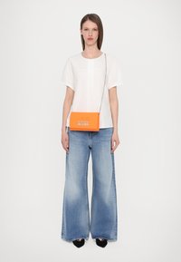 Orange crossbody bag with embossed "LOVE MOSCHINO," paired with a white short-sleeve top and wide-leg blue jeans. Black shoes complete the look.