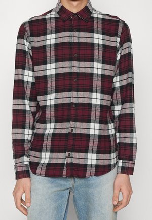 Man wearing buttoned-up long-sleeve shirt with red, black, gray, and white plaid pattern and light blue jeans.