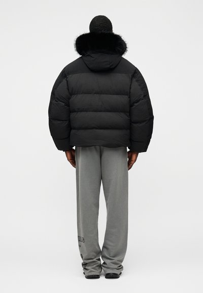 Casaco acolchoado preto com capuz forrado a pele, apresenta um corte quadrado e painéis horizontais acolchoados. Usado com calças de fato de treino cinzentas.