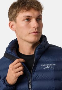 Giacca imbottita blu navy con chiusura a zip e colletto alto. Presenta un logo sul petto e una texture liscia e lucida.