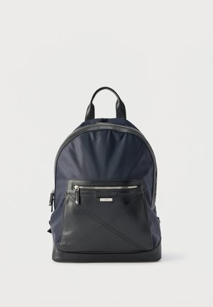 Zaino blu navy e nero con tasca frontale con cerniera, maniglia superiore e dettagli della cerniera argentati su sfondo bianco.