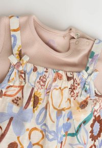 Mehka rožnato rebrasta enodelna obleka s pritrdilnimi sponkami, združena s kreamasto predpasnikom z rožnatim vzorcem, ki ima nastavljive trakove in pentljaste poudarke.