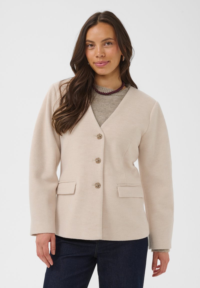 Femme aux longs cheveux bruns porte un blazer beige boutonné sur un haut gris, un jean foncé et un collier de perles, debout devant un fond blanc.