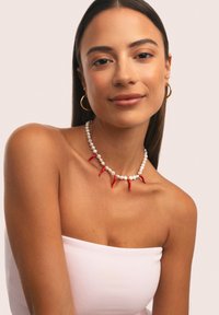 Donna con lunghi capelli scuri che indossa orecchini a cerchio dorati, una collana con peperoncini rossi e perle bianche, e un top bianco senza spalline su uno sfondo semplice.