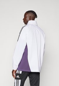 Hvid og lilla vindjakke med lange ærmer, høj krave og sorte detaljer. Har et sporty design med en løs pasform og elastiske manchetter.