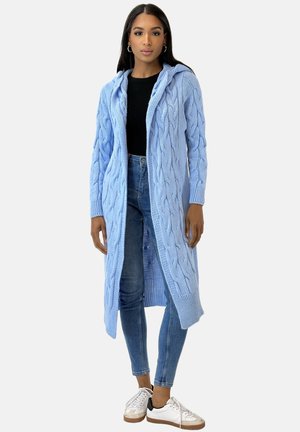 Lichtblauwe gebreide cardigan met een capuchon, voorzien van kabelpatronen en lange mouwen, gedragen over een zwarte top en blauwe jeans, gecombineerd met sneakers.