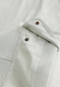 Tissu gris clair avec boutons-pression en métal, montrant des coutures détaillées et un rabat ouvert sur une poche de vêtement.