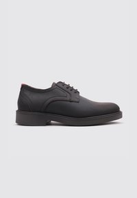 Zapato de cuero negro con una textura suave, punta redondeada y costuras marrones. Presenta un diseño con cordones y un acento rojo contrastante en el talón.