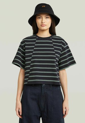 Jeune femme portant un haut court à manches courtes rayé noir et vert clair, un chapeau bob noir et un pantalon en jean foncé taille haute, debout devant un fond uni.