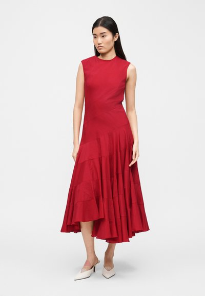 Γυναίκα με μακριά μαύρα μαλλιά, φορώντας ένα ασύμμετρο κόκκινο midi φόρεμα χωρίς μανίκια και λευκά μυτερά χαμηλά τακούνια, στέκεται μπροστά σε απλό φόντο.