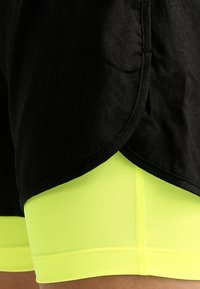 Pantalones cortos deportivos negros con acabado texturizado, superpuestos sobre pantalones cortos ajustados de color amarillo neón. La tela es suave y elástica con costuras visibles.