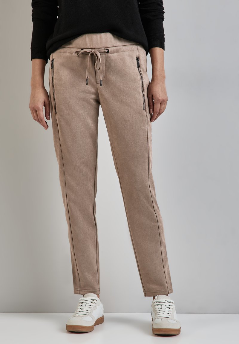 Beige tapered byxor i en strukturerad tyg, justerbar dragsko och zip-fickor i sidan, tillsammans med vita sneakers.