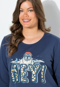 Námořnická košile s dlouhým rukávem, která zobrazuje grafiku lachtana s brýlemi a červenou čepicí, s textem "AHOJ!" v tučných, texturovaných písmenech.