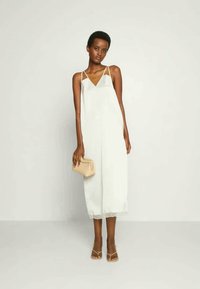 Λευκό ολόσωμο φόρεμα με τιράντες και λεπτομέρειες δαντέλας στο τελείωμα, διαθέτει χαλαρή εφαρμογή και σχέδιο v-λαιμό. Συνδυασμένο με μπεζ τσάντα και σандάλια.