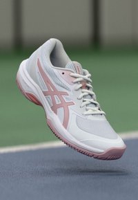 Hvit idrettssko med rosa detaljer, mesh- og syntetiske materialer, avrundet tå, teksturert såle og detaljert logo på siden.