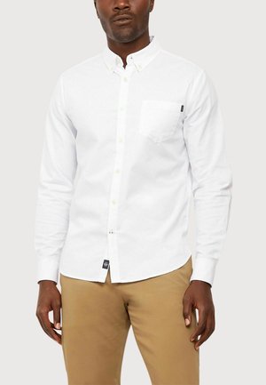 Camisa blanca de manga larga con botones y un bolsillo en el pecho, de tela de algodón, combinada con pantalones caqui.