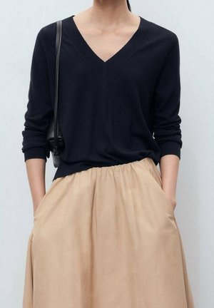 Personne portant un pull noir à col en V et une jupe beige, les mains dans les poches, portant un sac à bandoulière noir sur un fond clair uni.