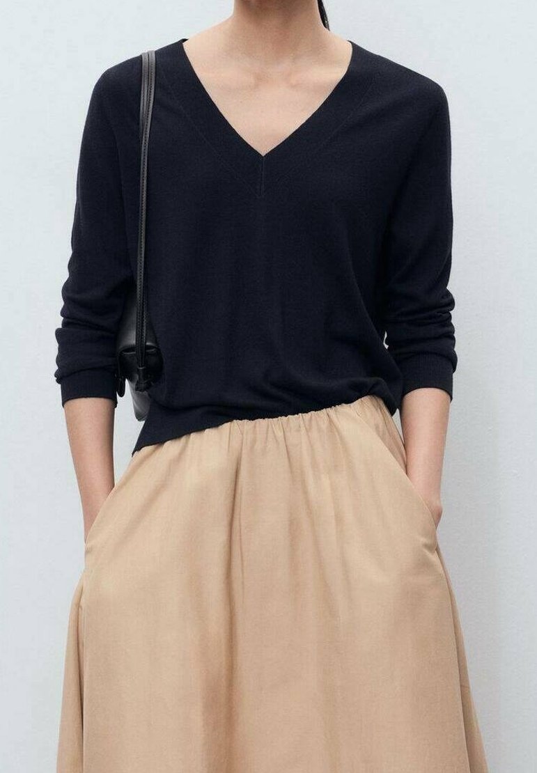 Personne portant un pull noir à col en V et une jupe beige, les mains dans les poches, portant un sac à bandoulière noir sur un fond clair uni.