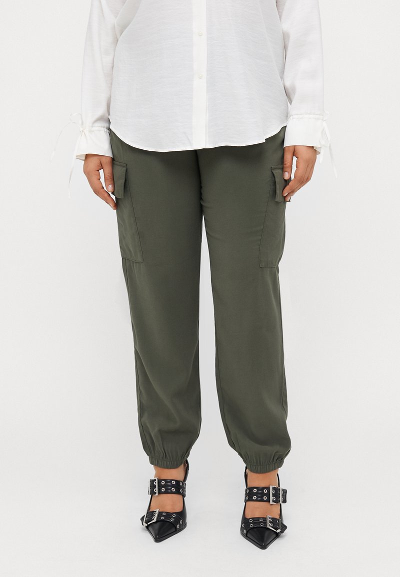 Ελαιόχρωμο cargo παντελόνι με ελαστικά μανίκια και πλευρικές τσέπες, σε συνδυασμό με μαύρα μυτερά παπούτσια με κοτσες. Λευκό πουκάμισο με κουμπιά.