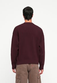 Pull en ribbed bordeaux avec une coupe décontractée, mettant en avant des épaules tombantes et un ourlet large. Associé à un pantalon cargo beige affichant des poches.