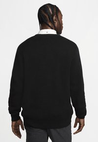 Μαύρο πλεκτό πουλόβερ με στρογγυλή λαιμόκοψη και μακριά μανίκια. Διαθέτει υφή στον ύφασμα και ριγωτές μανσέτες και στρίφωμα. Φοριέται πάνω από ένα λευκό πουκάμισο με γιακά.