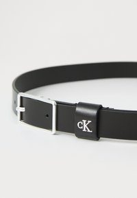 Juodas odinis diržas su sidabriniu stačiakampiu sagtimi ir odiniu kilpos elementu, įspaustu sidabriniu "cK" logotipu ant balto fono.