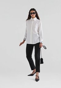 White button-up shirt with long sleeves, black accents on cuffs, paired with black tailored pants and black pointed-toe flats, holding a small bag.