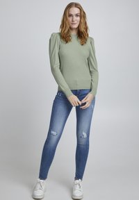 Hellgrüner ribbeliger Strickpullover mit Puffärmeln, kombiniert mit eng anliegenden, verwaschenen blauen Jeans und weißen Turnschuhen.