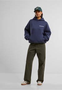 Marineblå hettegenser med lomme foran og hvitt logo, kombinert med løstsittende olivengrønne bukser og beige sneakers. Grønn caps som aksent.