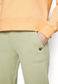 Meruňková mikina s žebrovanými manžetami, v kombinaci se zelenými tepláky s malou nášivkou srdce. Obě jsou vyrobeny z měkké, pohodlné tkaniny.