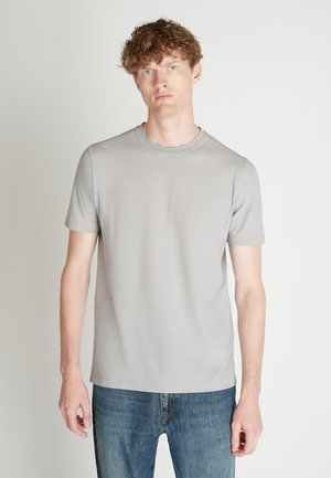 Junger Mann mit lockigem Haar, der ein schlichtes hellgraues T-Shirt mit kurzen Ärmeln und blaue Jeans trägt, steht vor einem weißen Hintergrund.