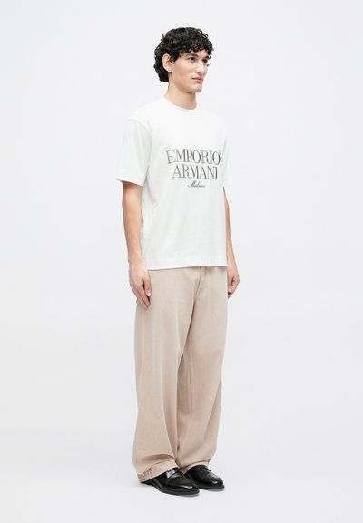 Biele bavlnené tričko s čiernym potlačeným nápisom „EMPORIO ARMANI Milano“, doplnené béžovými širokými nohavicami a čiernymi topánkami.