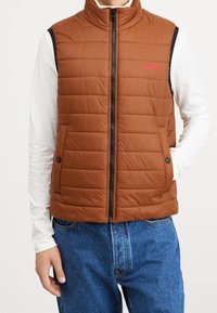 Braune gesteppte Weste mit hohem Kragen, Reißverschluss vorne, Seitentaschen und rotem Logo. Wird über einem weißen Langarmshirt und blauen Jeans getragen.