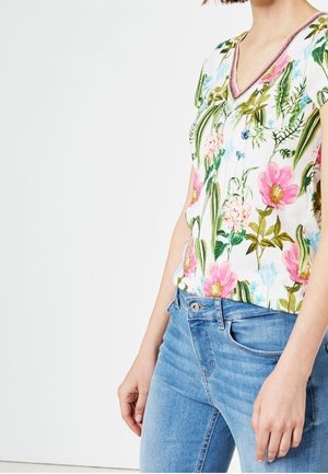 Mujer con blusa blanca de escote en V con estampado floral colorido, combinada con jeans azules de mezclilla, de pie contra un fondo claro y liso.