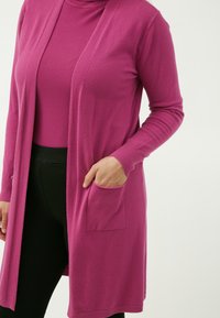 Cardigan lungo fucsia con apertura frontale, polsini a coste e una tasca frontale, abbinato a un dolcevita coordinato e pantaloni neri. Texture morbida.