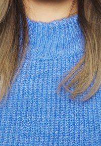Pull bleu tricoté avec une texture côtelée et un col rond, doté d'un tissu doux de poids moyen et de détails de couture visibles.