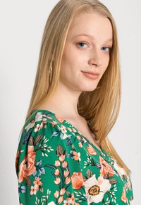 Blusa verde floral com decote em V profundo, apresentando flores laranja e brancas, detalhes de folhas e uma textura de tecido suave. Brincos de argola dourados visíveis.