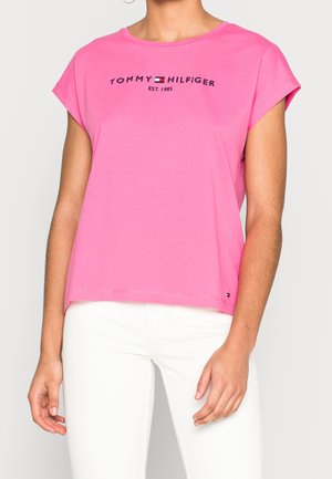 Kobieta w jaskraworóżowej koszulce Tommy Hilfiger z logo oraz białych spodniach, stojąca na tle jednolitego jasnego tła.