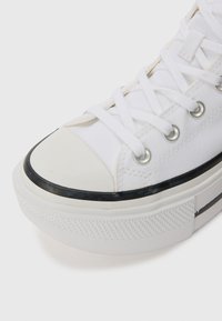 White canvas sneaker with a rounded toe, rubber toe cap, black rubber sole, silver eyelets, and flat white laces. Textured sole detail.