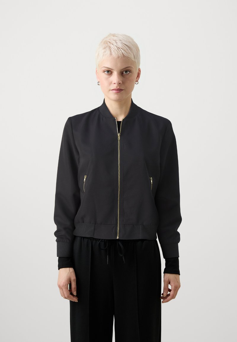 Giacca bomber nera con zip frontale, polsini e vita a coste. Presenta due tasche laterali con zip dorate. Tessuto liscio con una consistenza morbida.