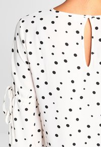 Blusa bianca con pois neri, apertura a chiave sulla schiena e maniche arricciate con lacci. Tessuto morbido, leggero e design casual.
