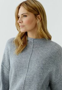 Vrouw met lang blond haar die een grijze gebreide trui draagt, nadenkend naar links kijkt tegen een effen lichte achtergrond.