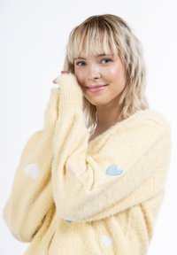Maglione giallo, morbido e peloso, con applicazioni di cuori pastello in rosa e blu sulle maniche. Tessuto soffice, design a V, vestibilità rilassata.